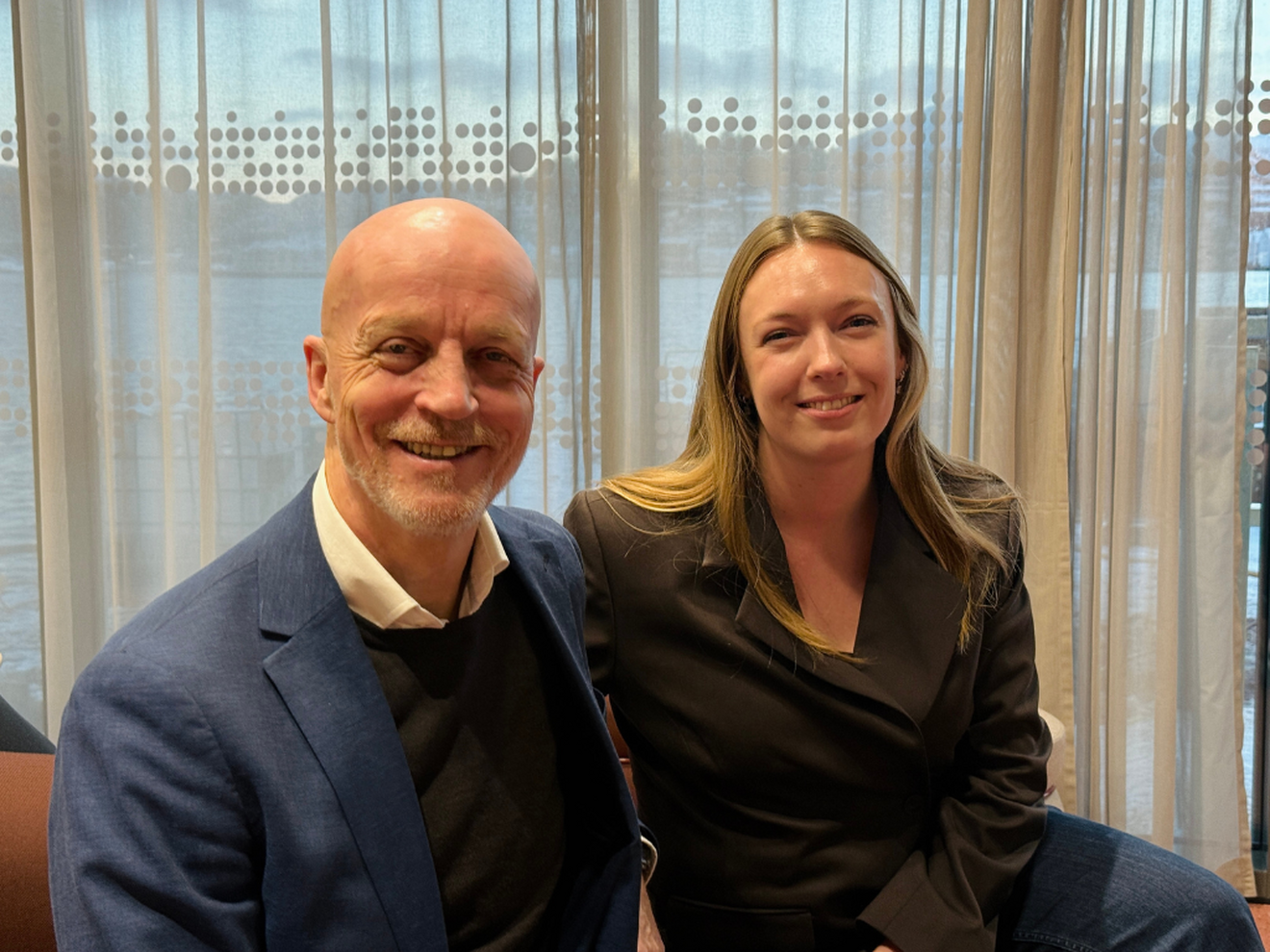 En mann og en kvinne sitter i ien orange sofa og smiler mot kamera. Mannen har blå dress, kvinnen brun. 