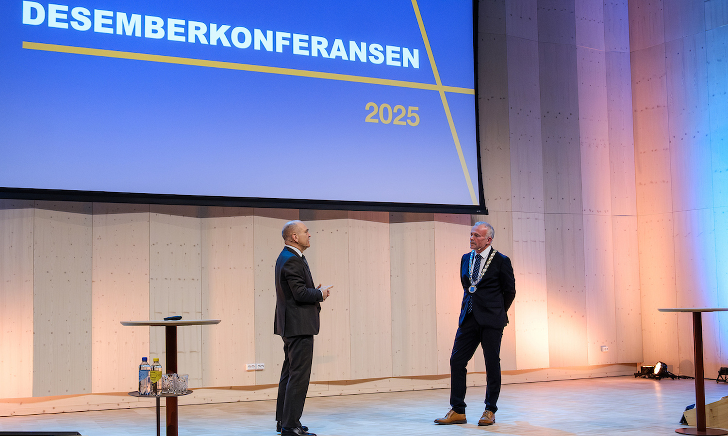 To menn i dress ser på hverandre fra et scenegulv. Bak dem skimtes påskriften 