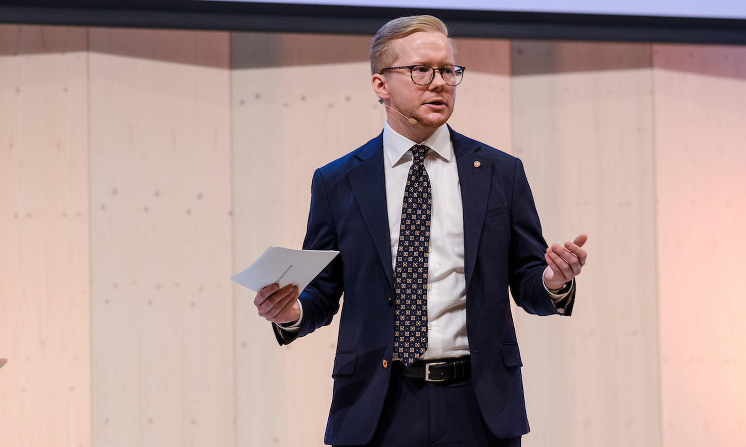 Mann i dress ser ut over publikum, og gestikulerer med armene mens han snakker