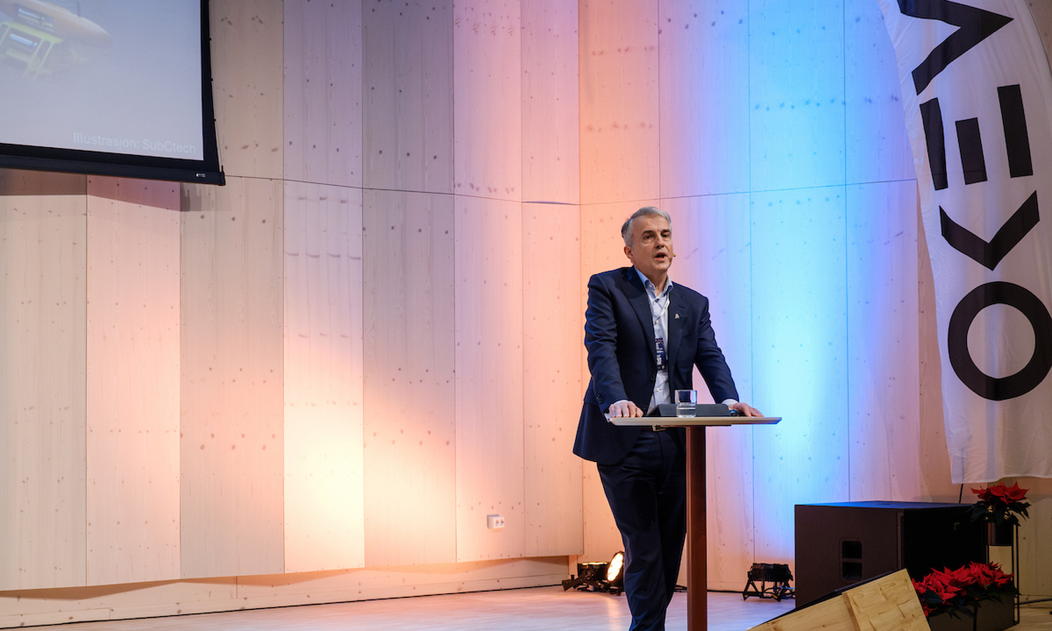 Mann i mørk dress og hvit skjorte holder i et rundt ståbord mens han snakker til publikum 