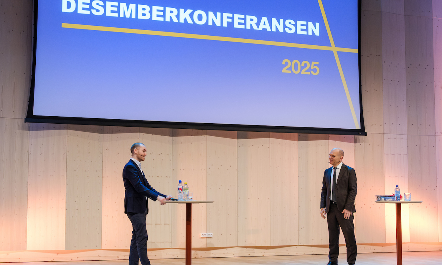 To menn i mørke dresser snakker sammen på et scenegulv av heltre. I bakgrunnen ser vi 