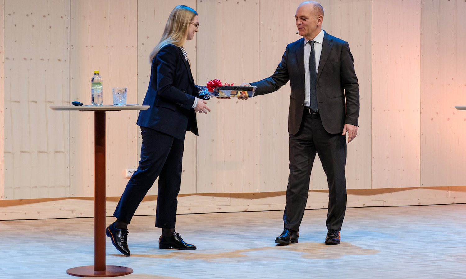 En kvinne i mørk dress mottar en sort pakke med rød sløyfe fra en mann i brun dress og hvit skjorte