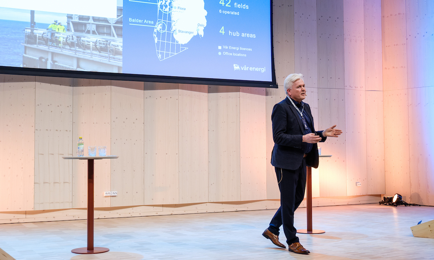 Mann i mørk dress, hvit skjorte og fyldig grått hår, gestikulerer mens han snakker. I bakgrunnen ser vi et bilde av hav, grå rør og to arbeidere i gule dresser 