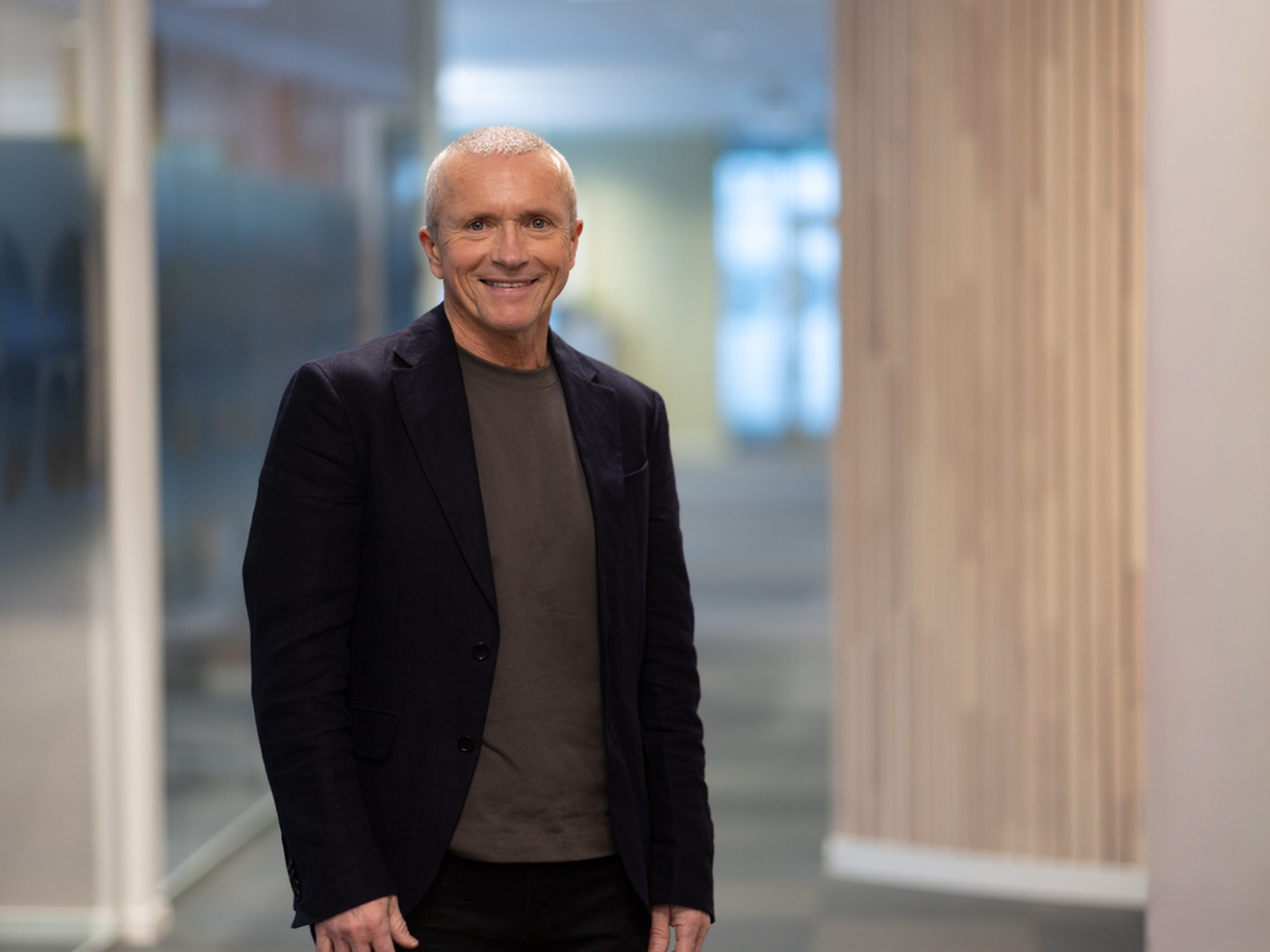 en mann med sort blazer, høyhalset grå genser smiler mot kamera. Bakgrunnen er uskarp og ser ut til å være i en korridor