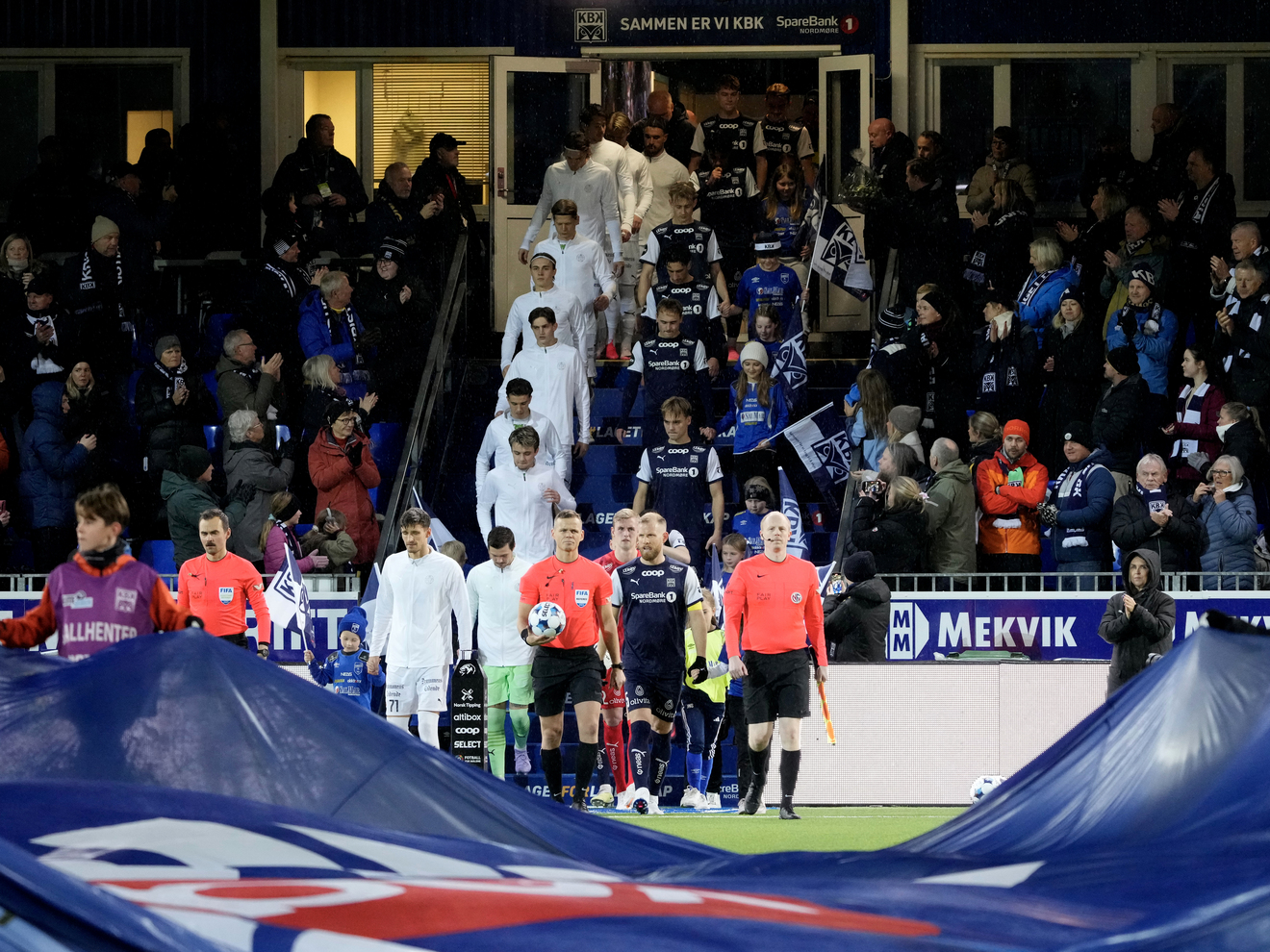 mange folk på en fotballbane, barn er på vei til å trekke en duk av banen mens fortballspillere i hvite drakter er på vei inn på banen