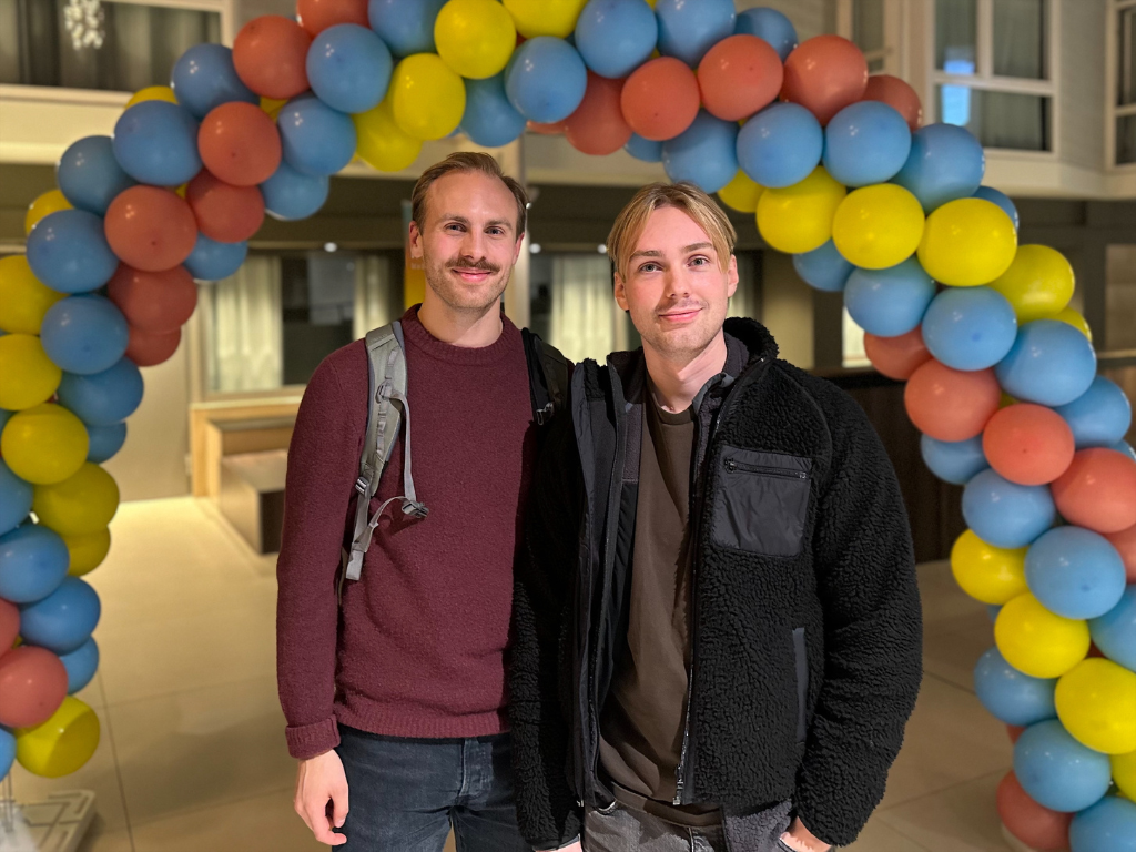 To unge menn står under en fargerik ballongbue og smiler mot kamera