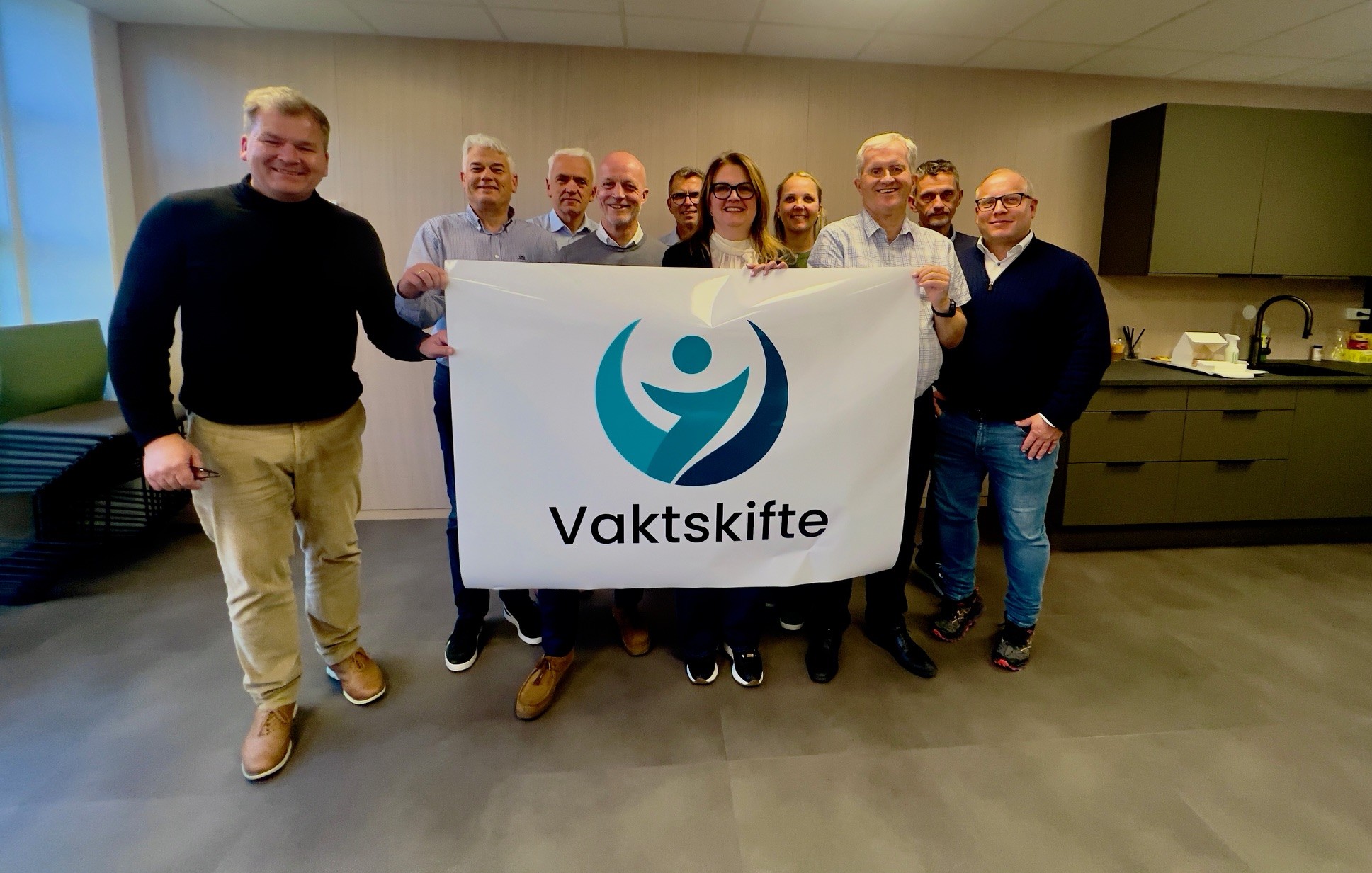 (F.V) , Henrik Dalelid Vedde (prosjektleder Vaktskifte), Erik Husby Pettersen (adm.dir. Varde), Ole Jonny Rugset (daglig leder, KNN), Heidi Nakken (adm.dir. Næringsforeningen i Ålesundsregionen), Idar Flø (styreleder, Hareid Næringsforum / representant for NæSS – næringsforeningene på Søre Sunnmøre), Marius Øksenvåg (adm.dir. Brisk AS) 10 mennesker står på et kjøkken med grønn innredning og holder opp en stor, hvit plakat der det står: Vaktskifte. Menneskene smiler mot kamera.
